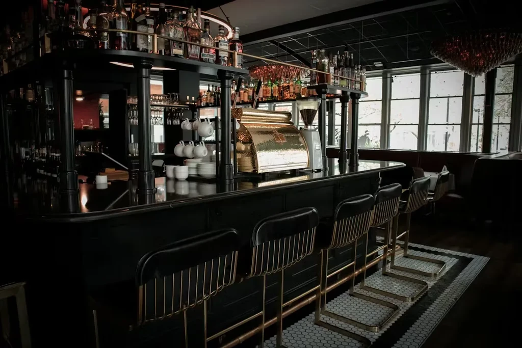 Black bar counter with brass stools, liquor bottles, and gold espresso machine.