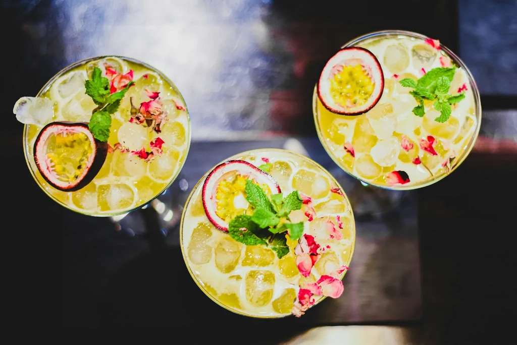Three pale yellow cocktails garnished with passion fruit, mint, and petals.