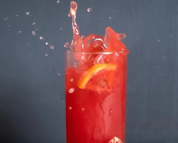 Red drink splashing from a clear glass, with a citrus slice.