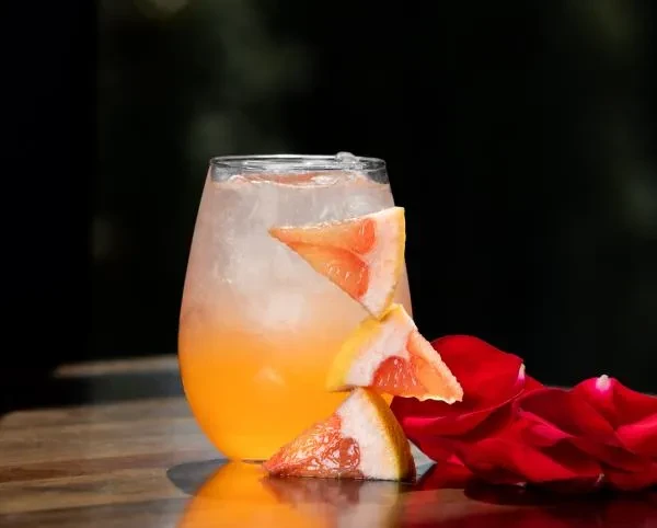 Two-toned drink with grapefruit, ice, and rose petals on wood.
