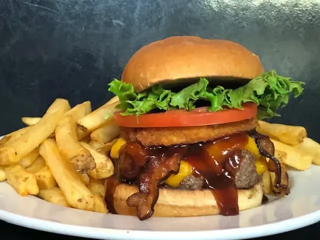 Close-up of a large burger with bacon, BBQ sauce, and golden fries.