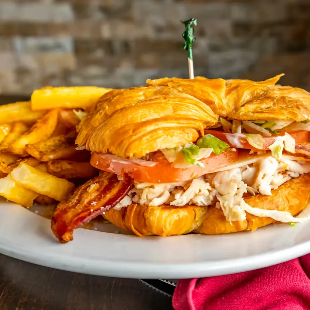 Flaky croissant club sandwich with bacon, lettuce, tomato, and fries.