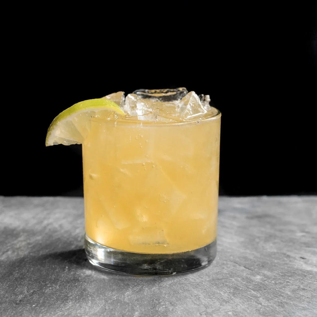 Amber drink with ice and lime in a tumbler on stone.