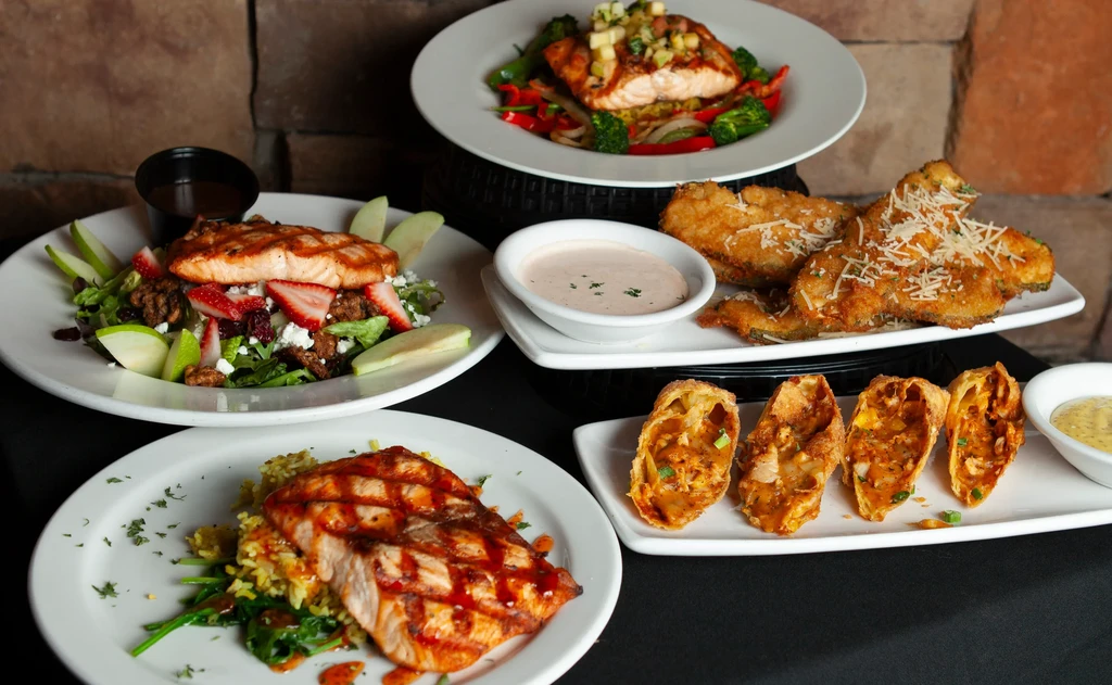 Grilled salmon dishes, salad, and fried appetizers on a dark table.