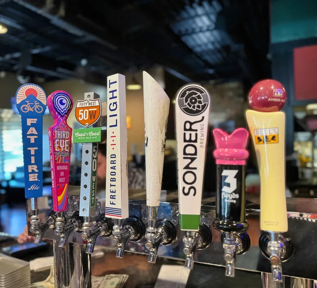 Row of nine varied beer tap handles with brand logos at a bar.