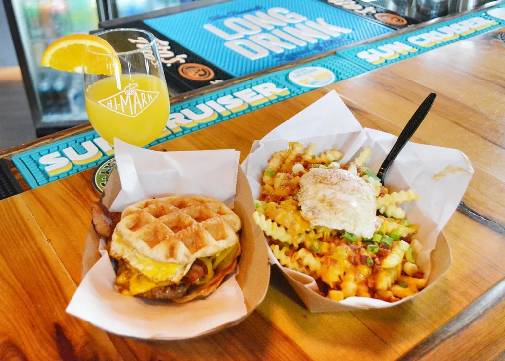 Mimosa, bacon-egg waffle sandwich, and loaded waffle fries on wooden bar.