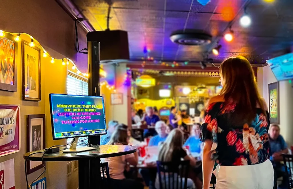 Woman watches karaoke monitor in a lively, blurred bar.