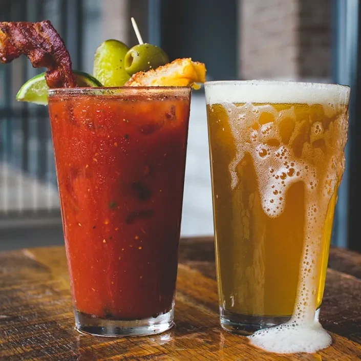 Elaborate Bloody Mary and overflowing beer on a rustic wooden table.
