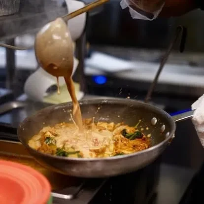 Hand pours creamy sauce into pan with noodles and green vegetables.