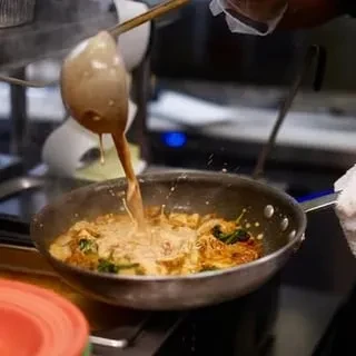 Chef's hand pours sauce into sauté pan with chicken and greens.