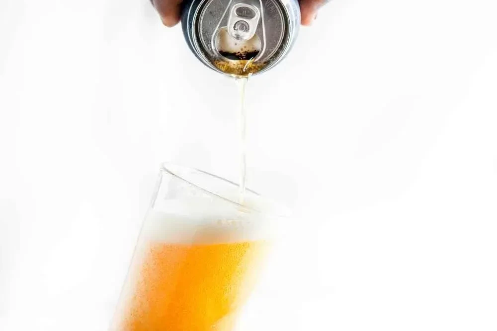Dark-skinned hand pours golden, effervescent drink from can into glass.