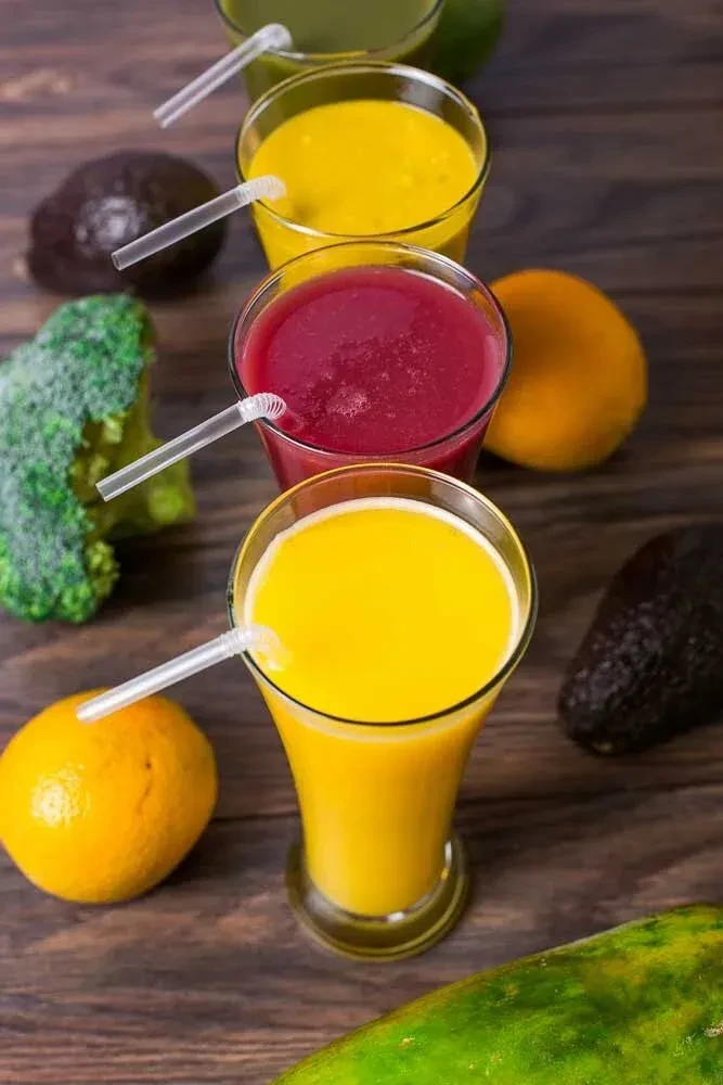 Colorful smoothies in glasses with straws, surrounded by fresh fruits and vegetables.