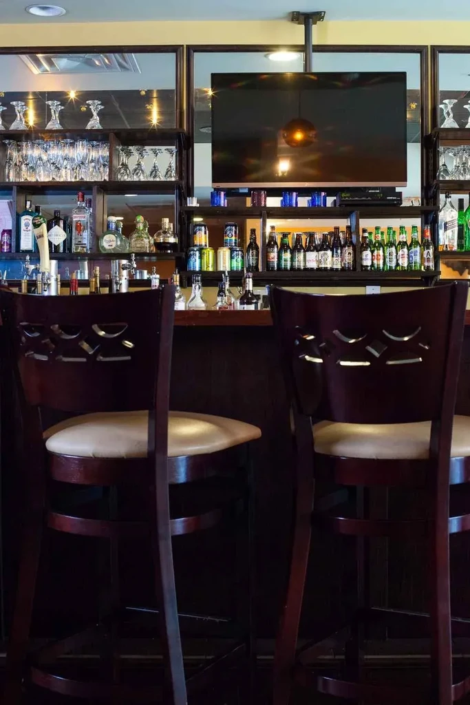 Warmly lit, well-stocked bar with bottles, glassware, stools, and a TV.