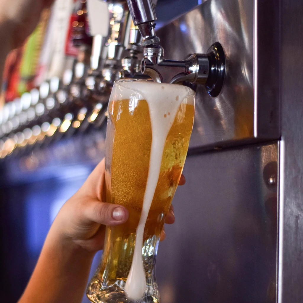 Hand holds glass of golden beer overflowing with foam from a tap.