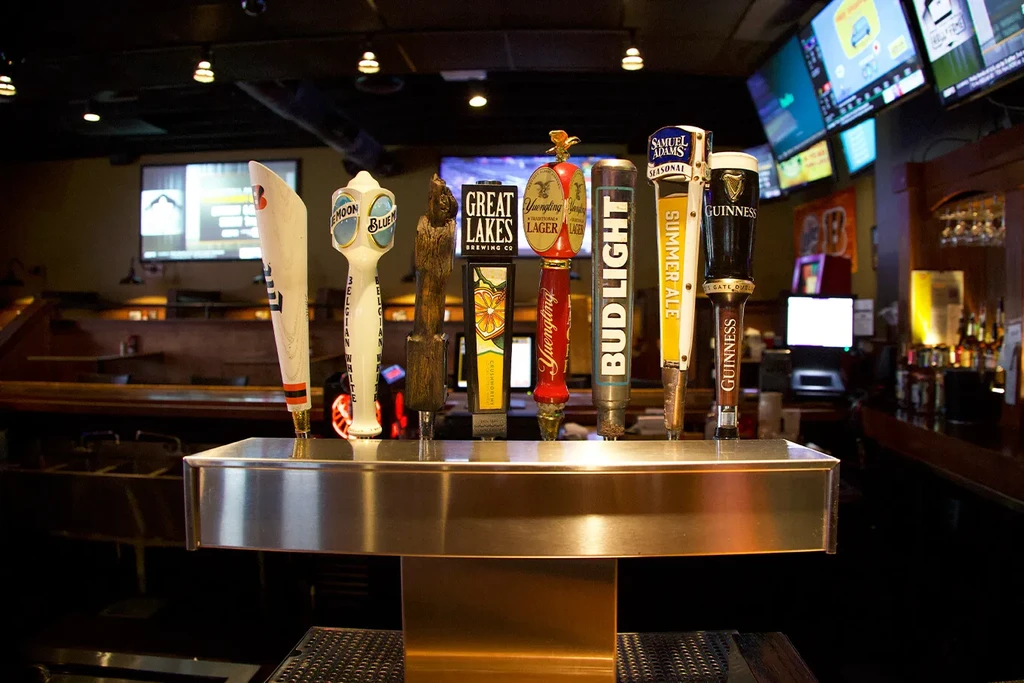Diverse beer tap handles on a stainless steel tower in a sports bar.