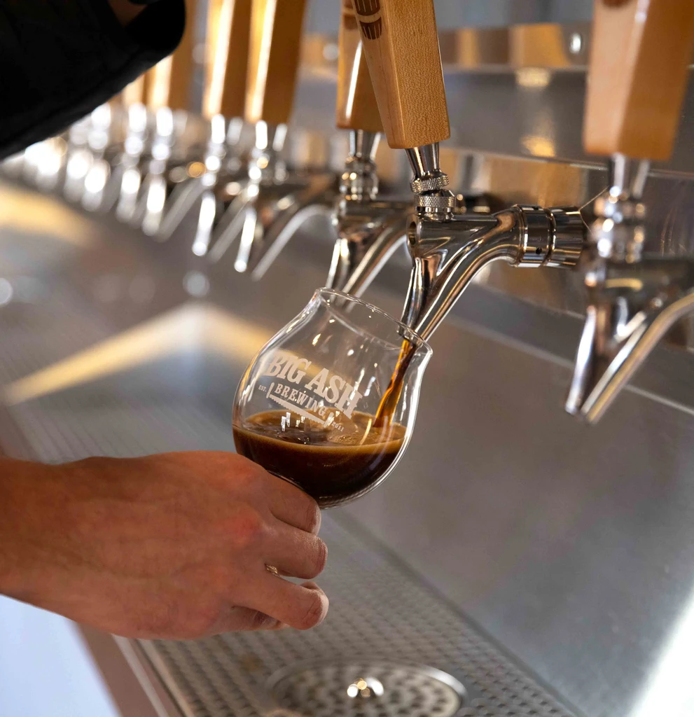 Hand pouring dark beer into a "BIG ASH BREWING" glass from a tap.