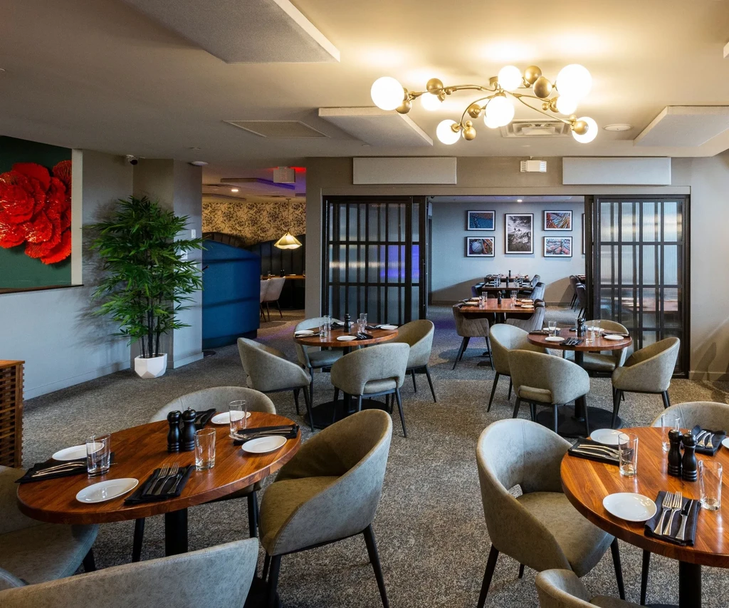 Empty modern restaurant with wooden tables, grey chairs, and brass chandelier.