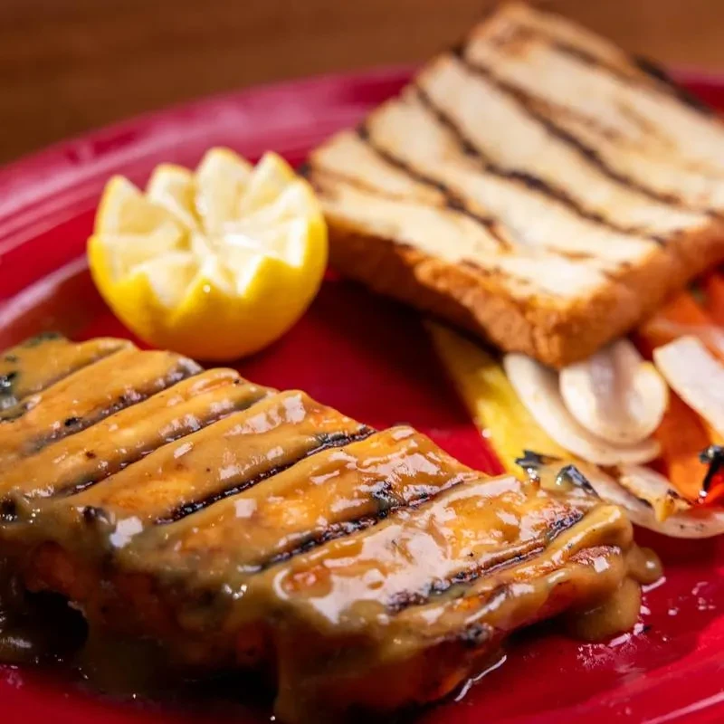Grilled food with sauce, lemon, bread, and vegetables on red plate.