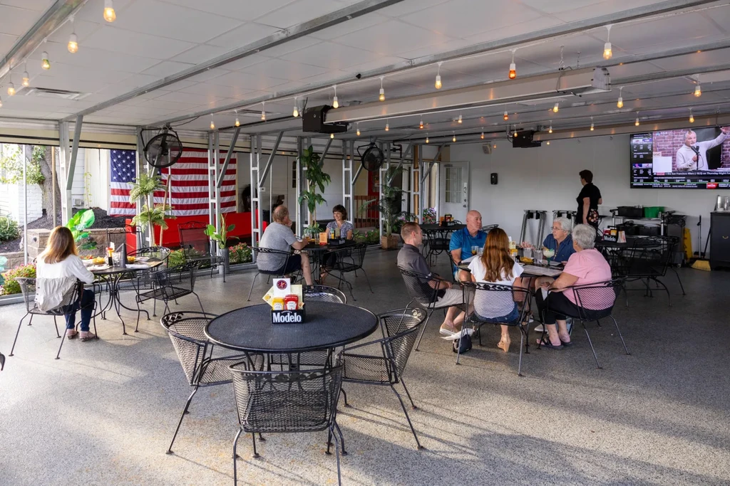 Lively patio scene with patrons, string lights, US flag, and television.