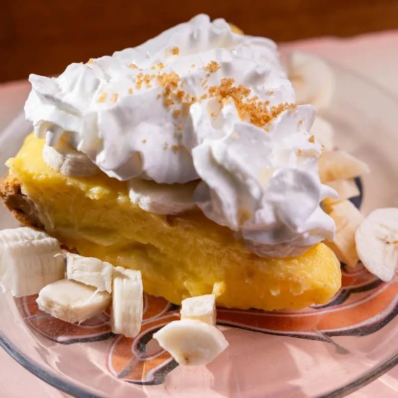 Golden banana cream pie slice, whipped cream, crumbs, and banana slices.