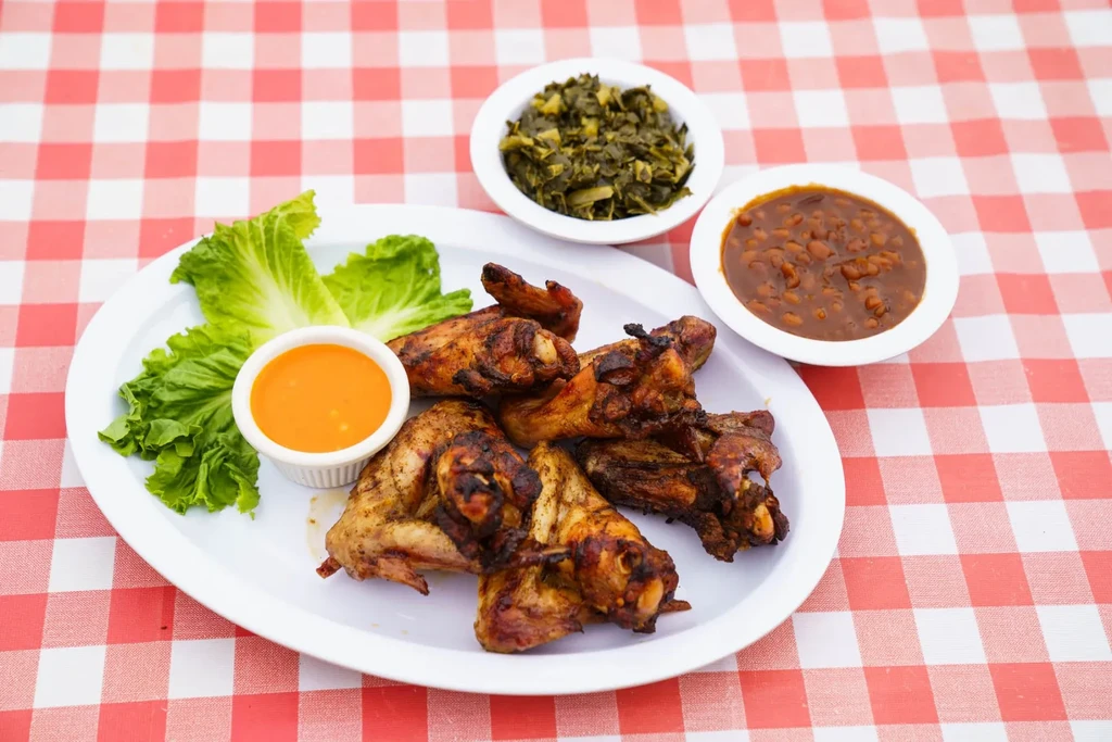 Grilled chicken wings, collard greens, baked beans, and sauce on checkered tablecloth.