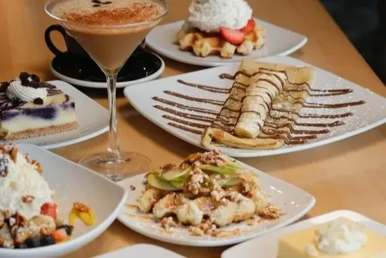 Varied dessert spread: crepes, waffles, cakes, and a creamy cocktail on wood.