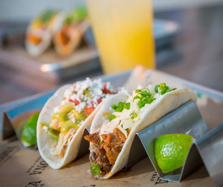 Two tacos on a holder with meat, cheese, sauce, and lime.