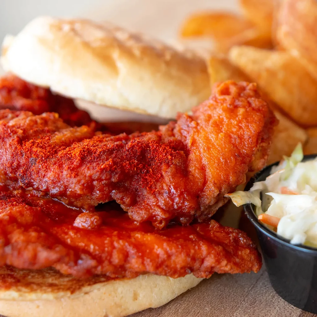 Close-up spicy fried chicken sandwich, coleslaw, and fries on wooden surface.