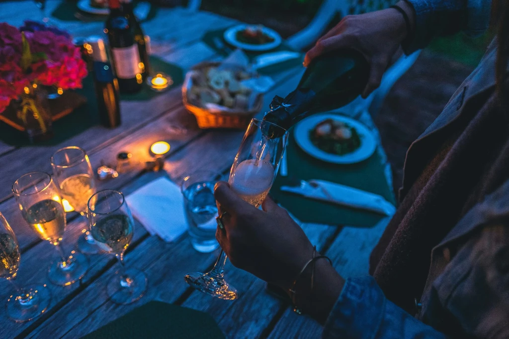 Close-up of hand pouring sparkling wine into flute on candlelit rustic table.