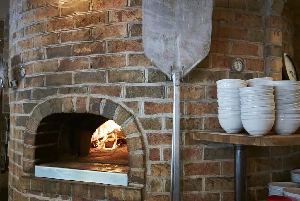 Rustic brick oven with fire, pizza peel, and ceramic bowls.