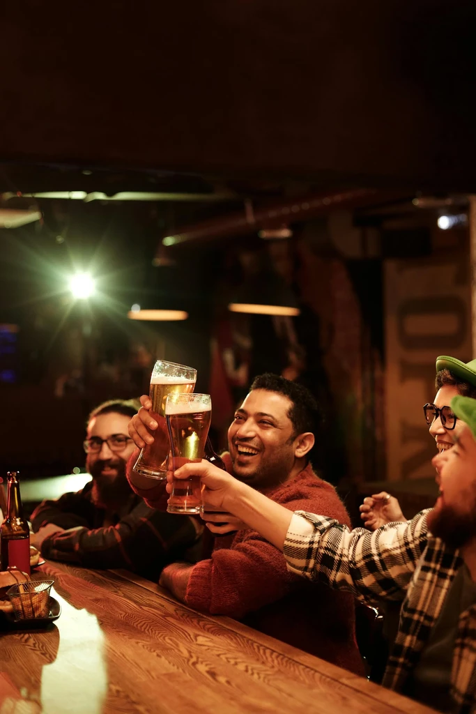 Smiling men toast beer glasses at a warm, dimly lit bar.