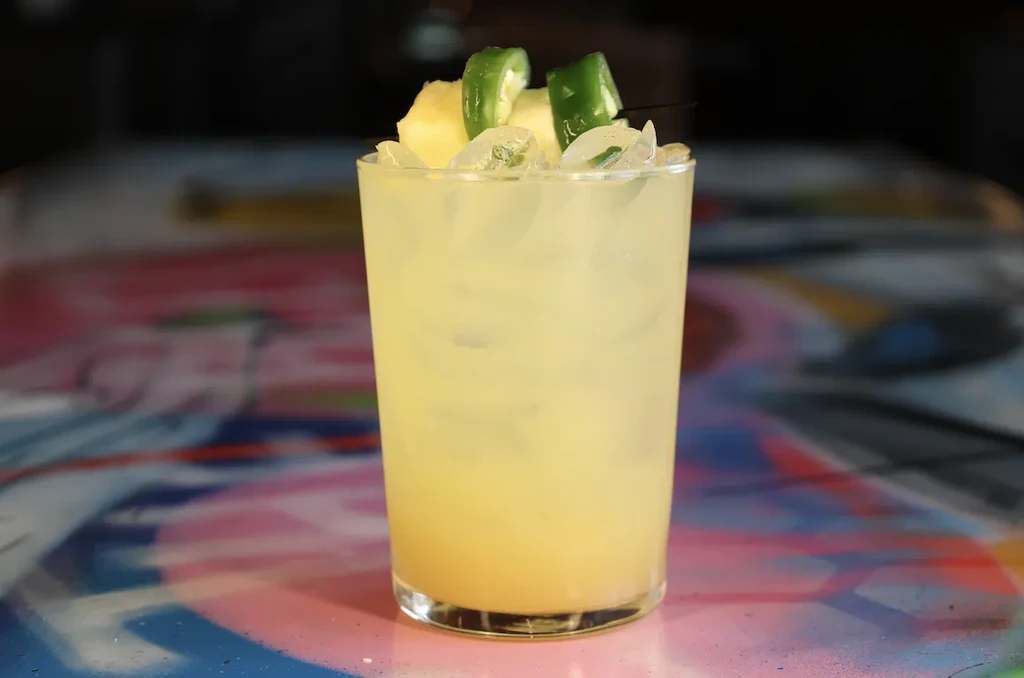 Pale yellow cocktail with pineapple and chili garnish on abstract table.