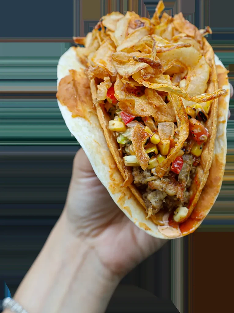 Hand holds a large taco with meat, corn, peppers, crispy onions.