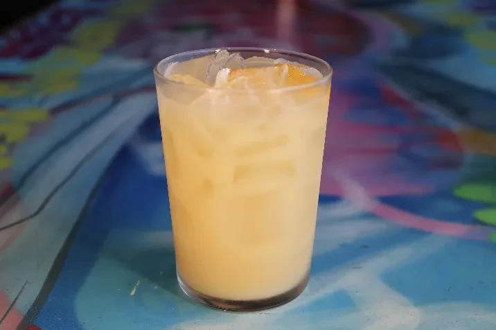 Tall glass of pale yellow drink with ice and citrus garnish.