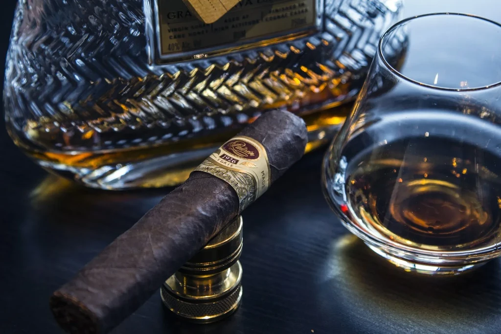 Dark still life: unlit cigar, whiskey glass, and "GRAN" spirit bottle.