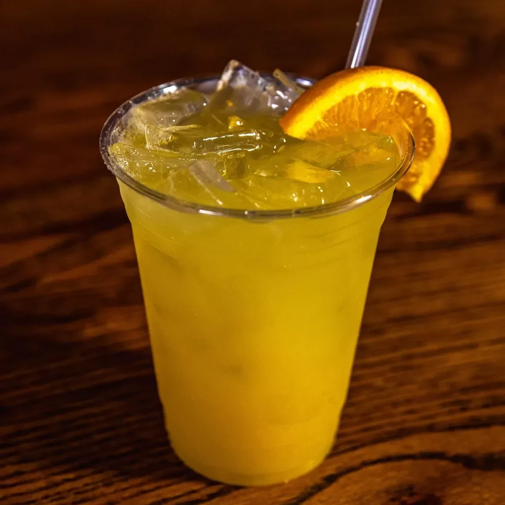 Yellow-orange iced drink in plastic cup with orange slice, straw, wood.