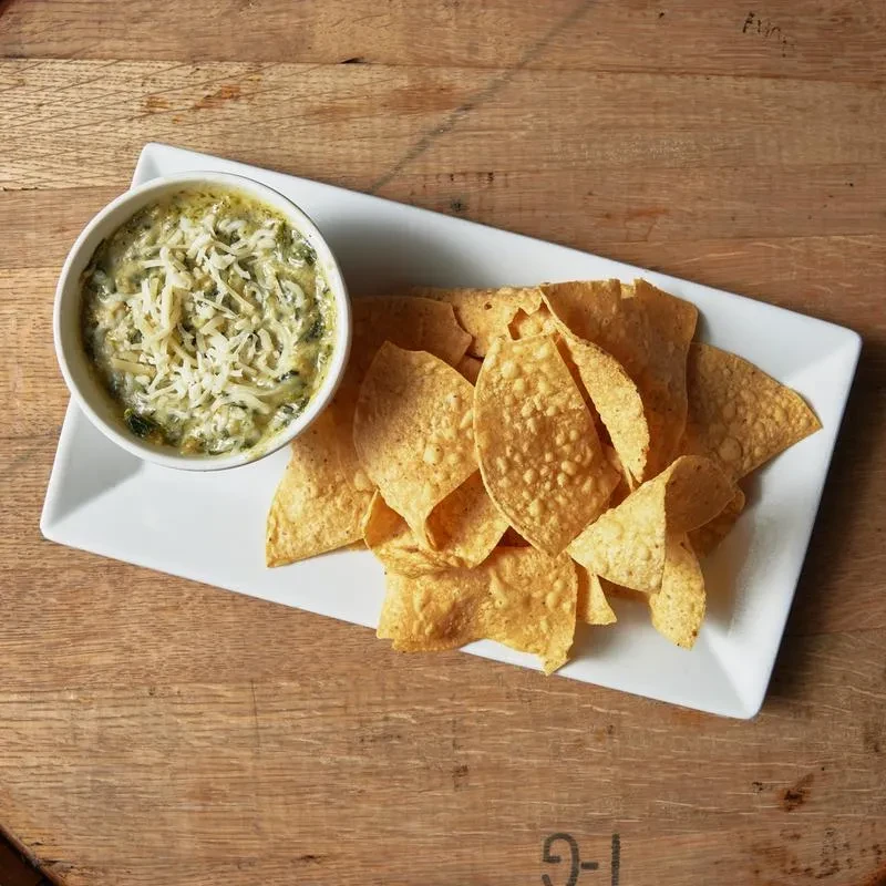 Spinach dip topped with cheese, served with tortilla chips on wood.