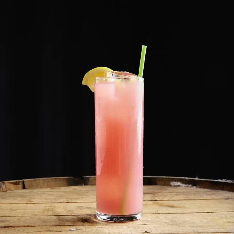 Pink drink in tall glass with ice, green straw, citrus, wood.