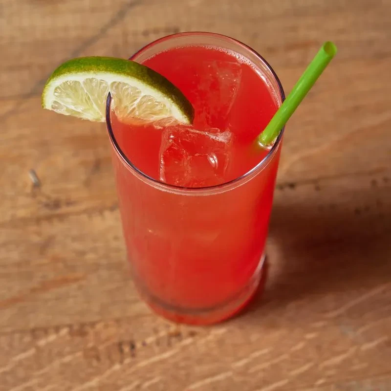 Red-orange drink with ice, lime, and green straw on a table.