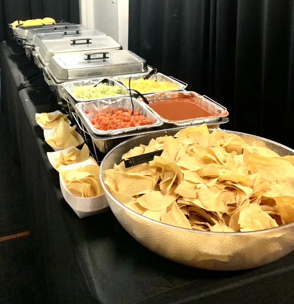 Buffet table with tortilla chips, cheese, lettuce, tomatoes, and salsa.