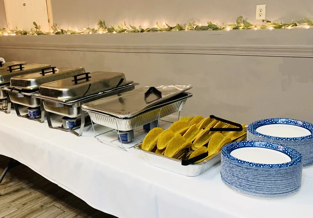 Buffet table with chafing dishes, taco shells, plates, and festive garland.