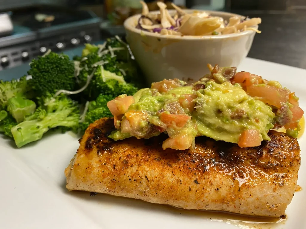 Pan-seared fish with avocado salsa, cheesy broccoli on a white plate.