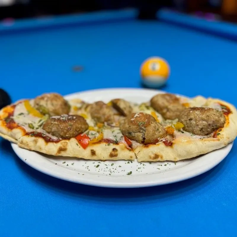Meatball pizza on a plate, with a blurred pool table background.