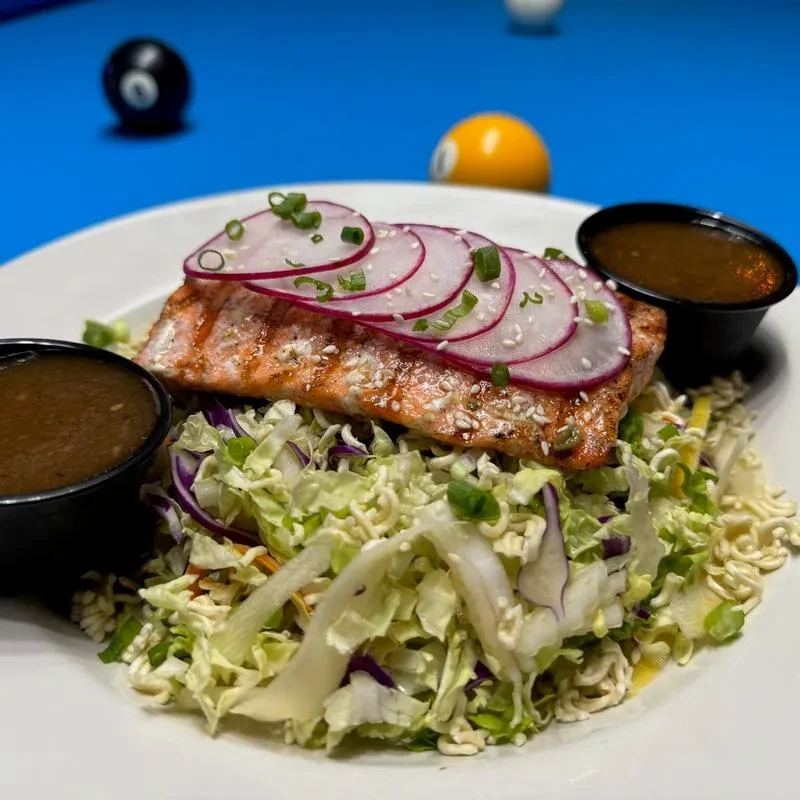 Grilled salmon fillet with radishes, slaw, and sauce, on a white plate.