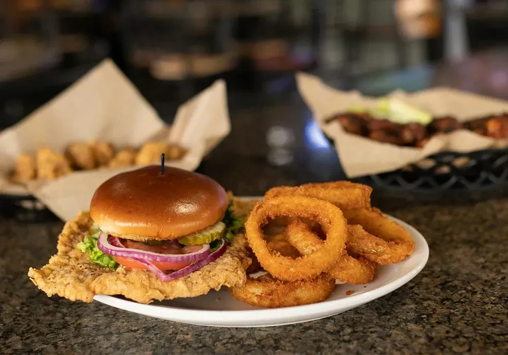 Fried cutlet sandwich with lettuce, tomato, and onion rings on white plate.