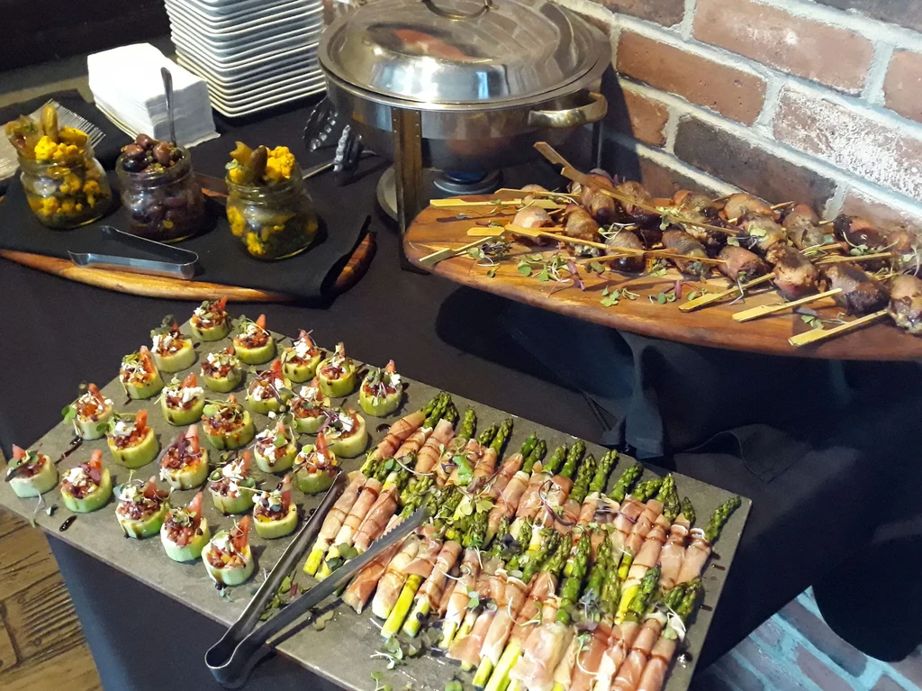 Buffet display featuring prosciutto asparagus, cucumber canapés, pickled items, and skewers.