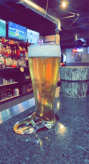 Boot-shaped glass of amber beer in a dimly lit bar setting.