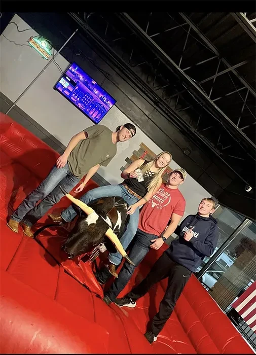 Four young adults smiling around a mechanical bull at a bar.