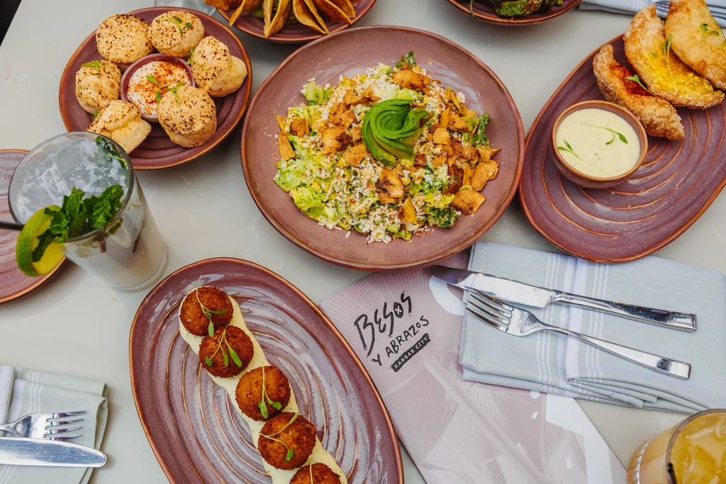 Flat lay dining table with diverse dishes, cocktail, and 'Besos y Abrazos' menu.