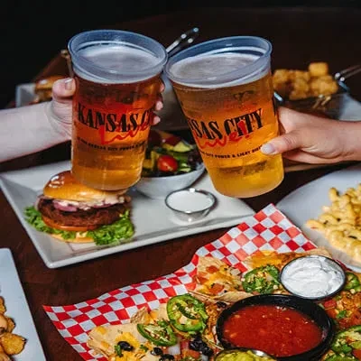 Hands clinking Kansas City beer cups above nachos and a burger.